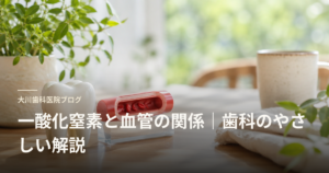 大川歯科医院ブログ 一酸化窒素と血管の関係 | 歯科のやさしい解説。歯の模型と赤色の血管モデルが木製テーブルに置かれ、観葉植物が背景のヘッダー画像。